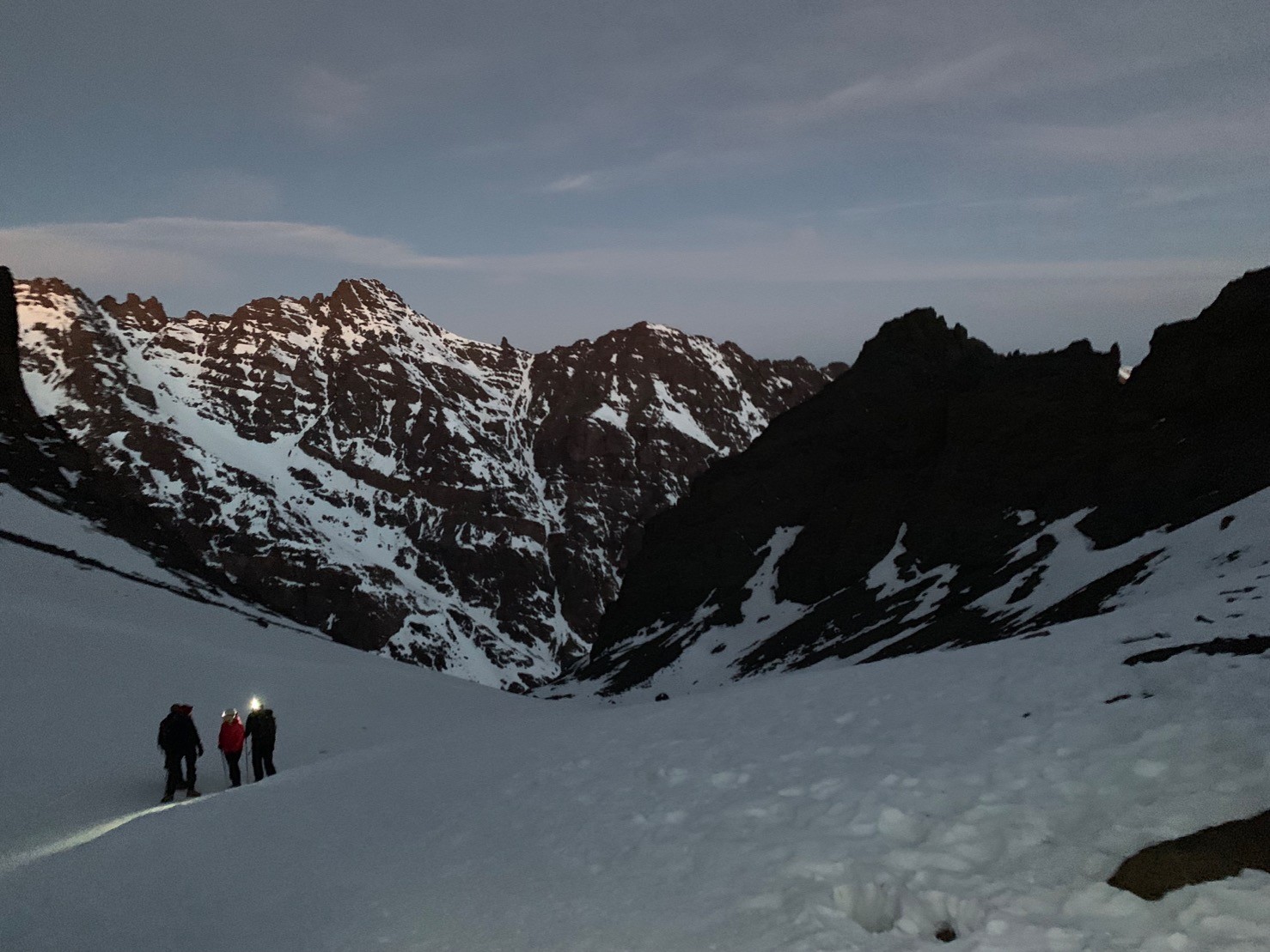 Several walkers wearing head torches. In the background are mountains and in the foreground there is snow.