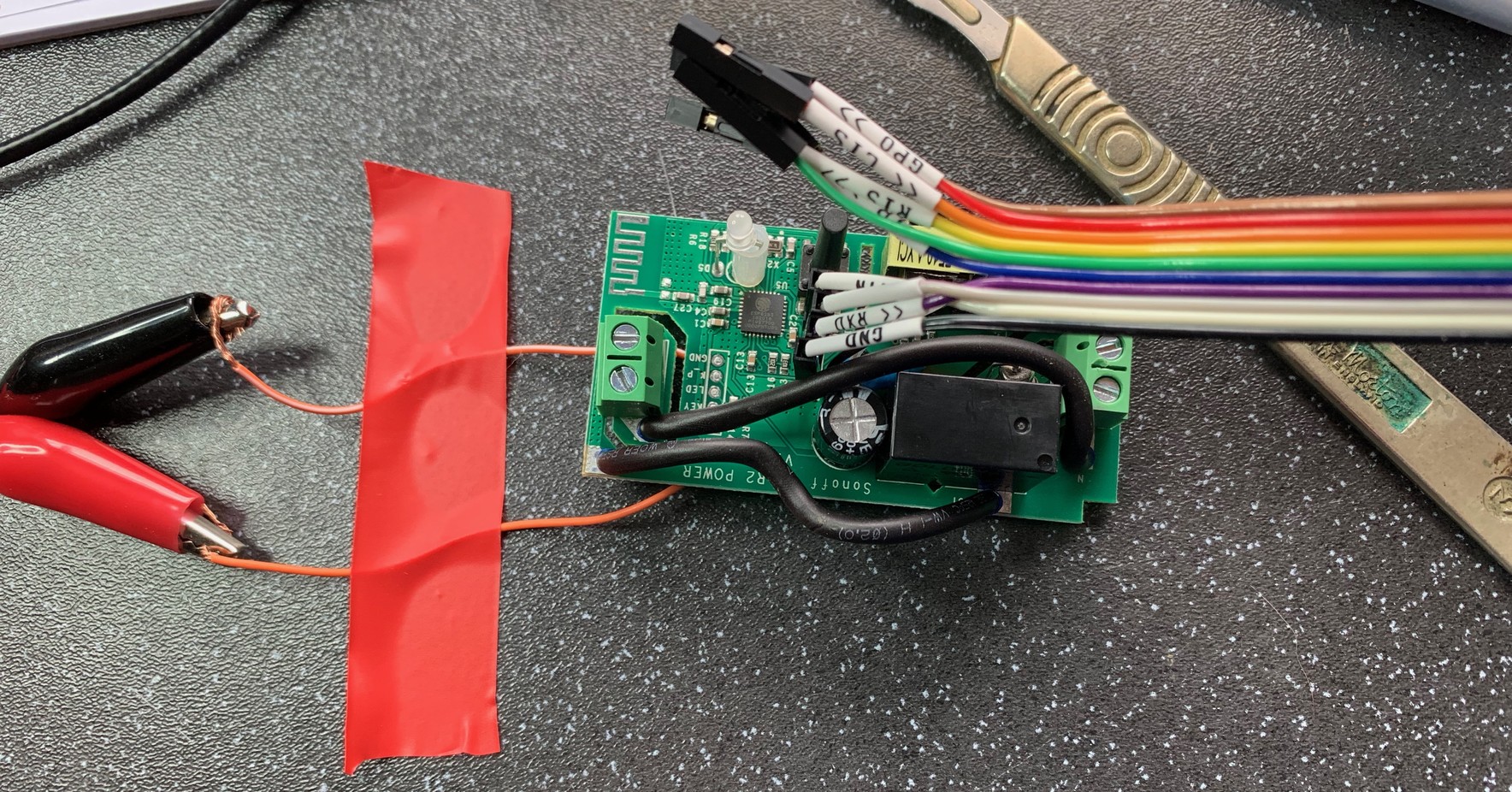 A small circuit board taped to a bench. On the left are two crocodile clips attached to two small wires which go under the board. These provide external power. Coming out of the centre of the board is a ribbon cable. This goes to the UART. The board is rectangular with a relay on the bottom right corner and small screw terminals at each end.