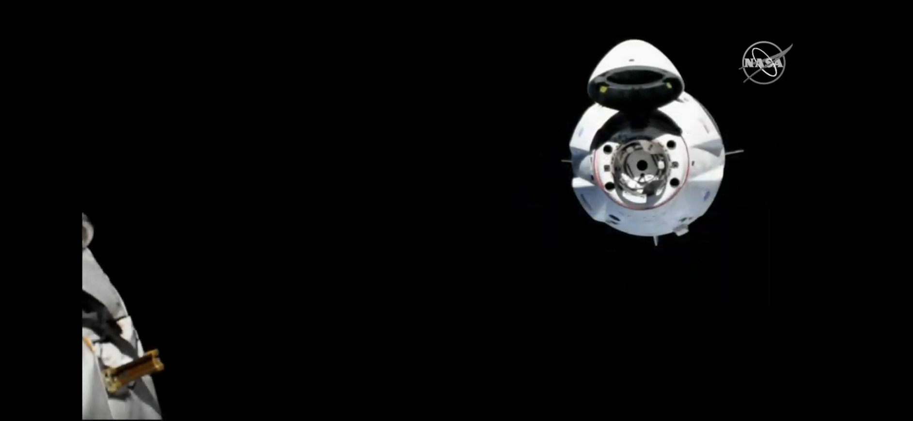 A view of the Dragon capsule from the ISS. The capture is a round white object in top right, and a small indistinct second of the ISS in in the bottom left. The capsule has a bulge on top, which is the open nose cone, and several small apertures in a ring, which are part of the docking system.