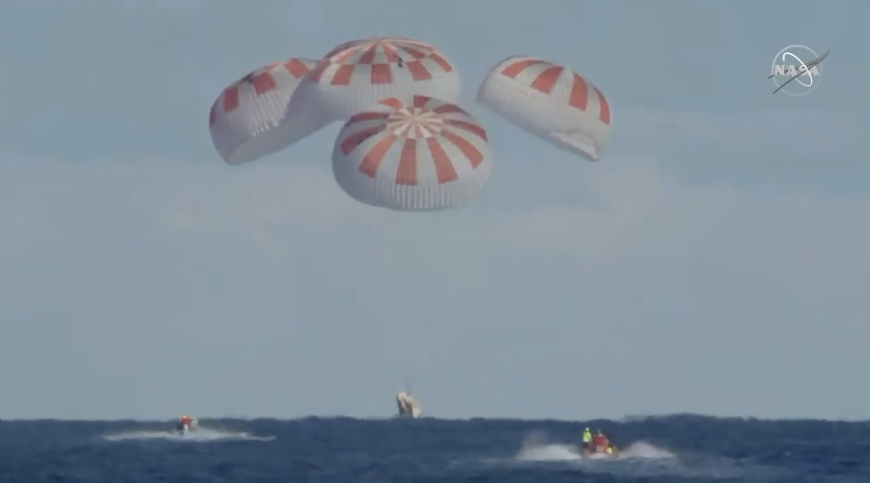 The Dragon 2 capsule landing in the Atlantic Ocean. There are four parachutes at the top of the image, the sea near the bottom, and the dragon capsule landing in the sea. There are two small boats heading towards the capsule.