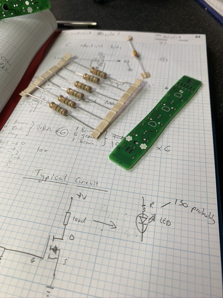 A squared note book with some large resistors and a narrow PCB on the top half of page. There are some notes and circuit diagrams on the bottom half.