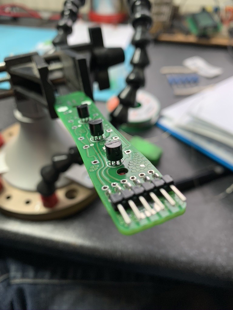 A PCB in a clamp. There are three MOSFETs in a row along the top of the board, and a 6 pin header close to the camera. 